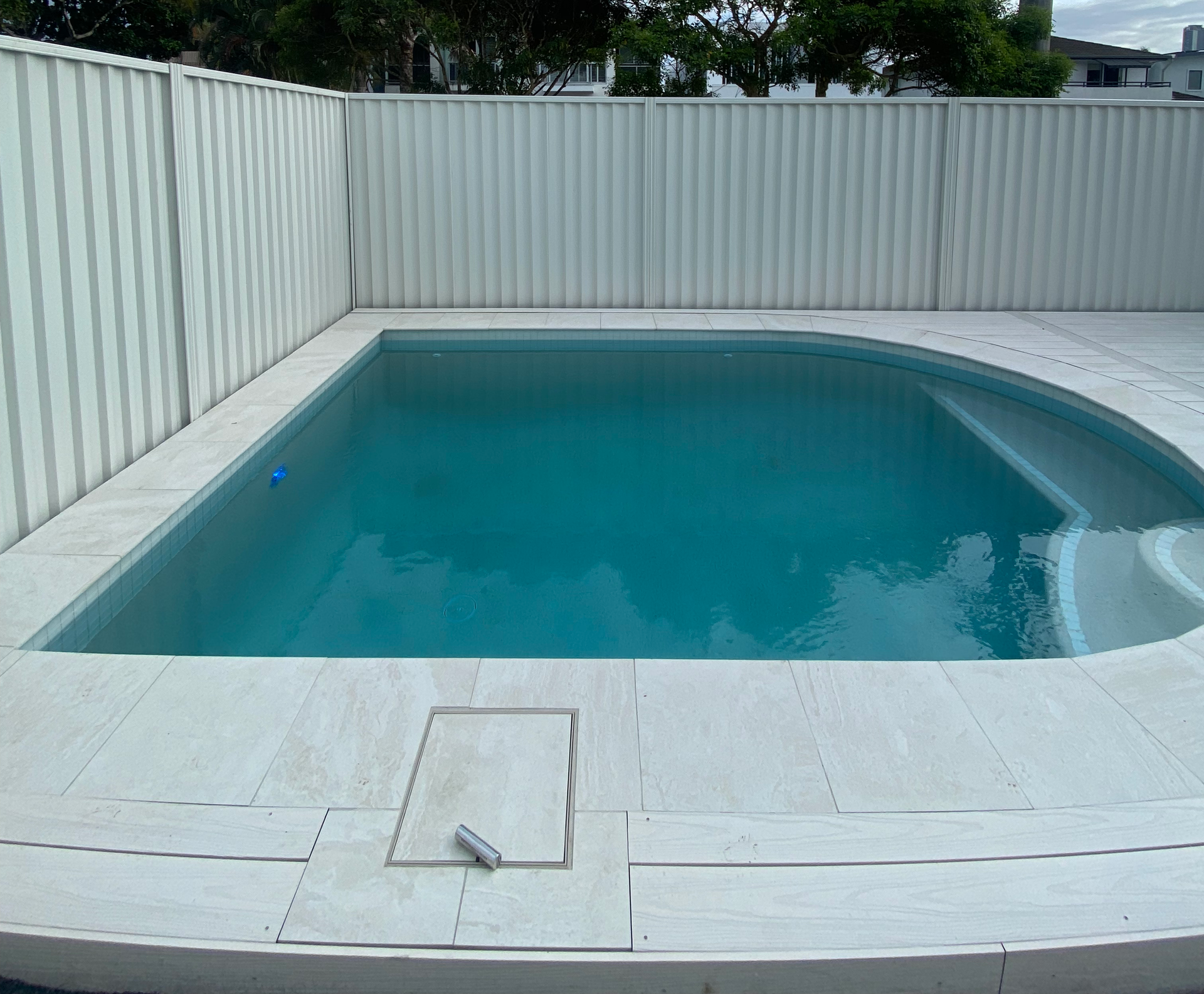 Small backyard swimming pool with clear blue water, white fence, and cloudy sky overhead