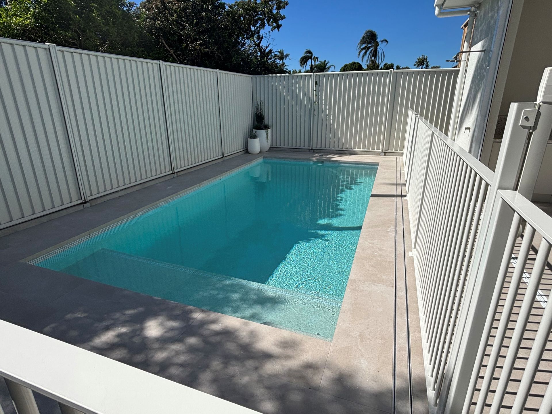 Rectangular backyard swimming pool with turquoise water, white fence, and shaded deck beside a house