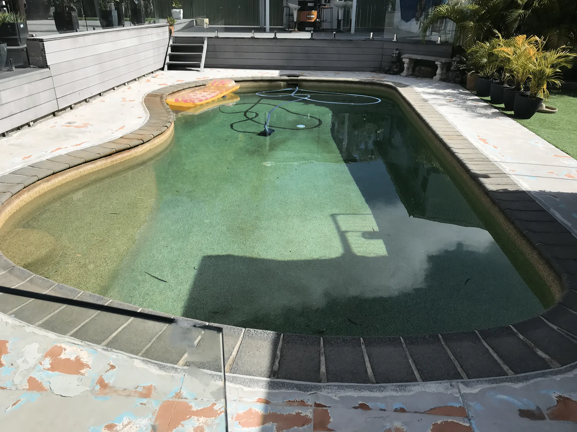 Small outdoor swimming pool with green water, tiled deck, and patio chairs in bright sunlight