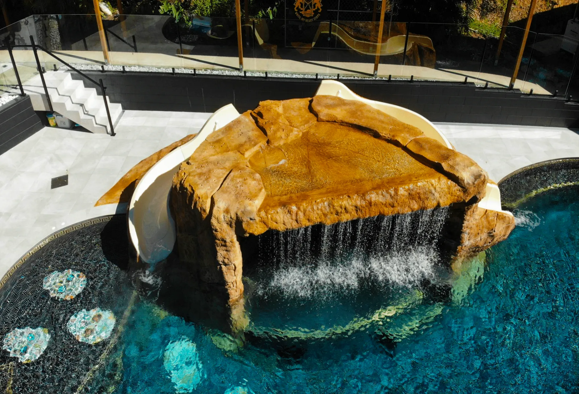 Waterfall spilling from a large tan rock into a blue pool at a sunny outdoor resort