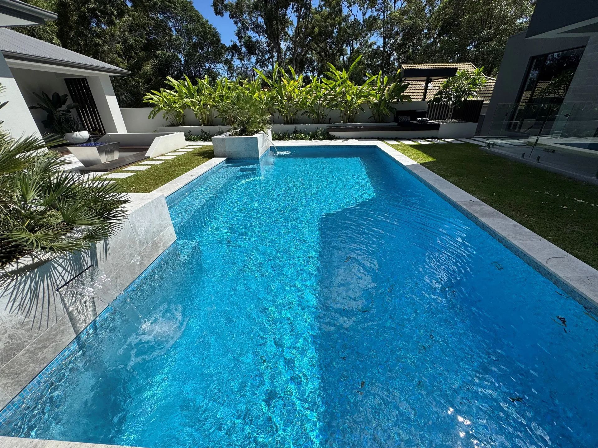 Modern backyard with a bright blue swimming pool, patio seating, and trees in sunlight
