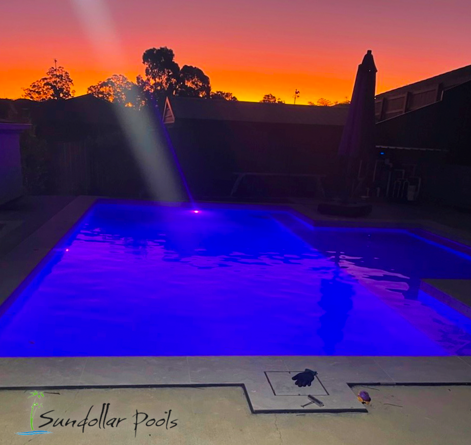 Backyard pool glowing blue at sunset, with orange sky and silhouetted trees.