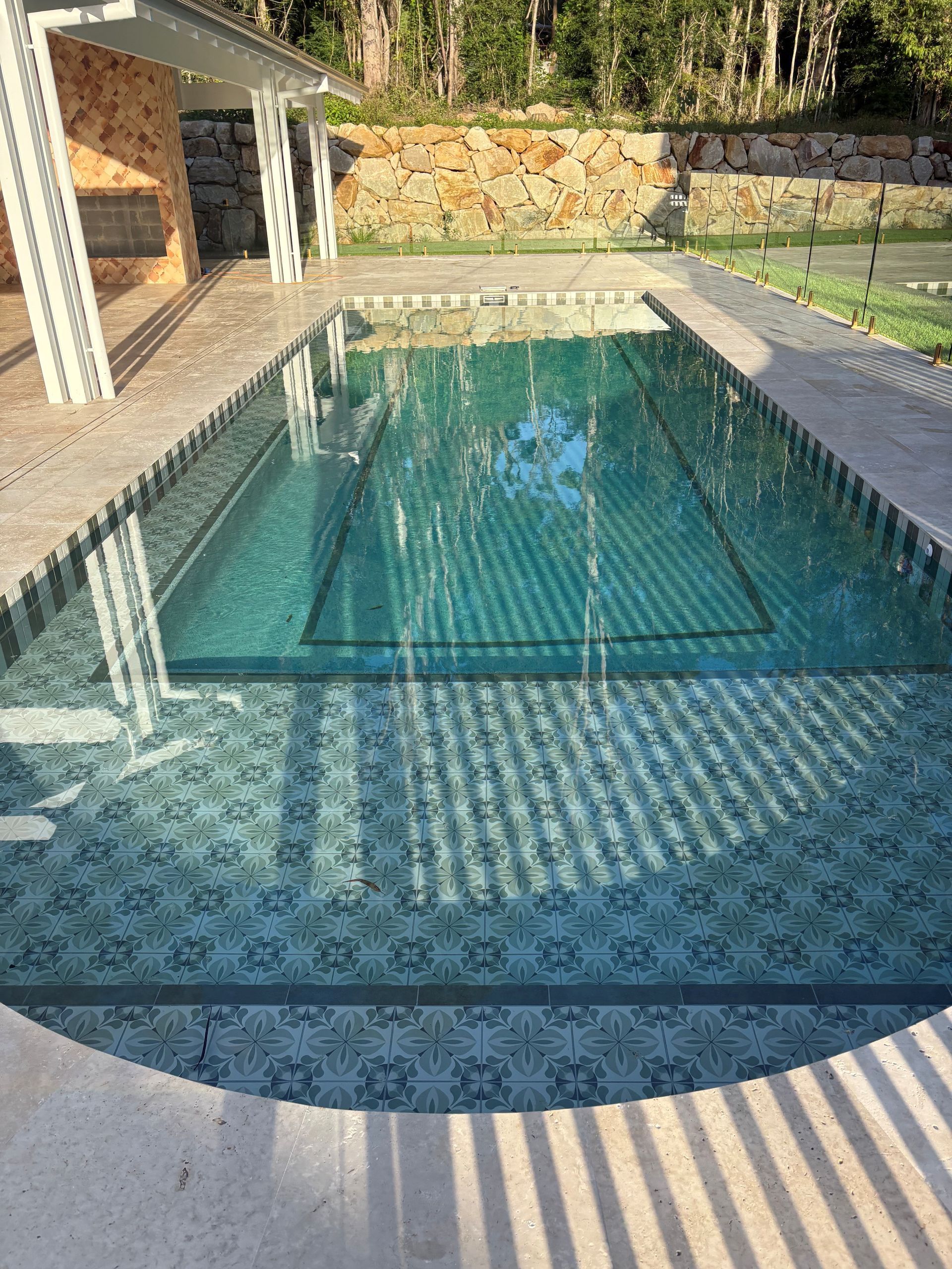 Outdoor swimming pool with striped sunlight reflections, blue water, and surrounding stone patio and greenery