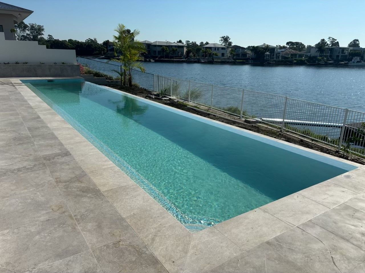 Long rectangular pool beside a lake, with blue water and a stone deck under a clear sky.