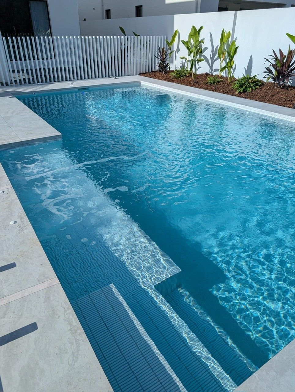 Blue backyard swimming pool with steps, white fence, and landscaped plants alongside the deck