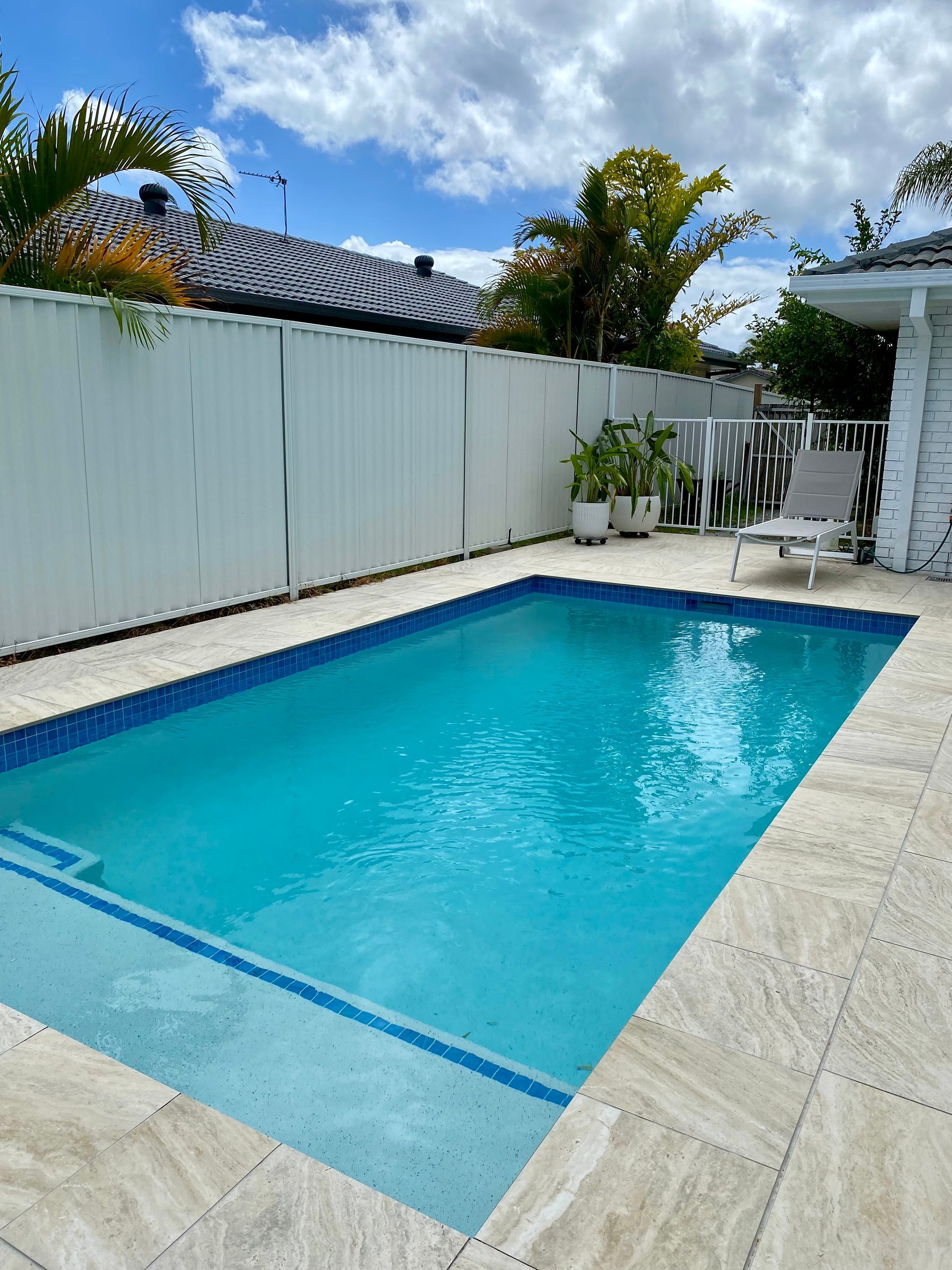 Small backyard swimming pool with white fence, stone deck, and tropical plants under a blue sky