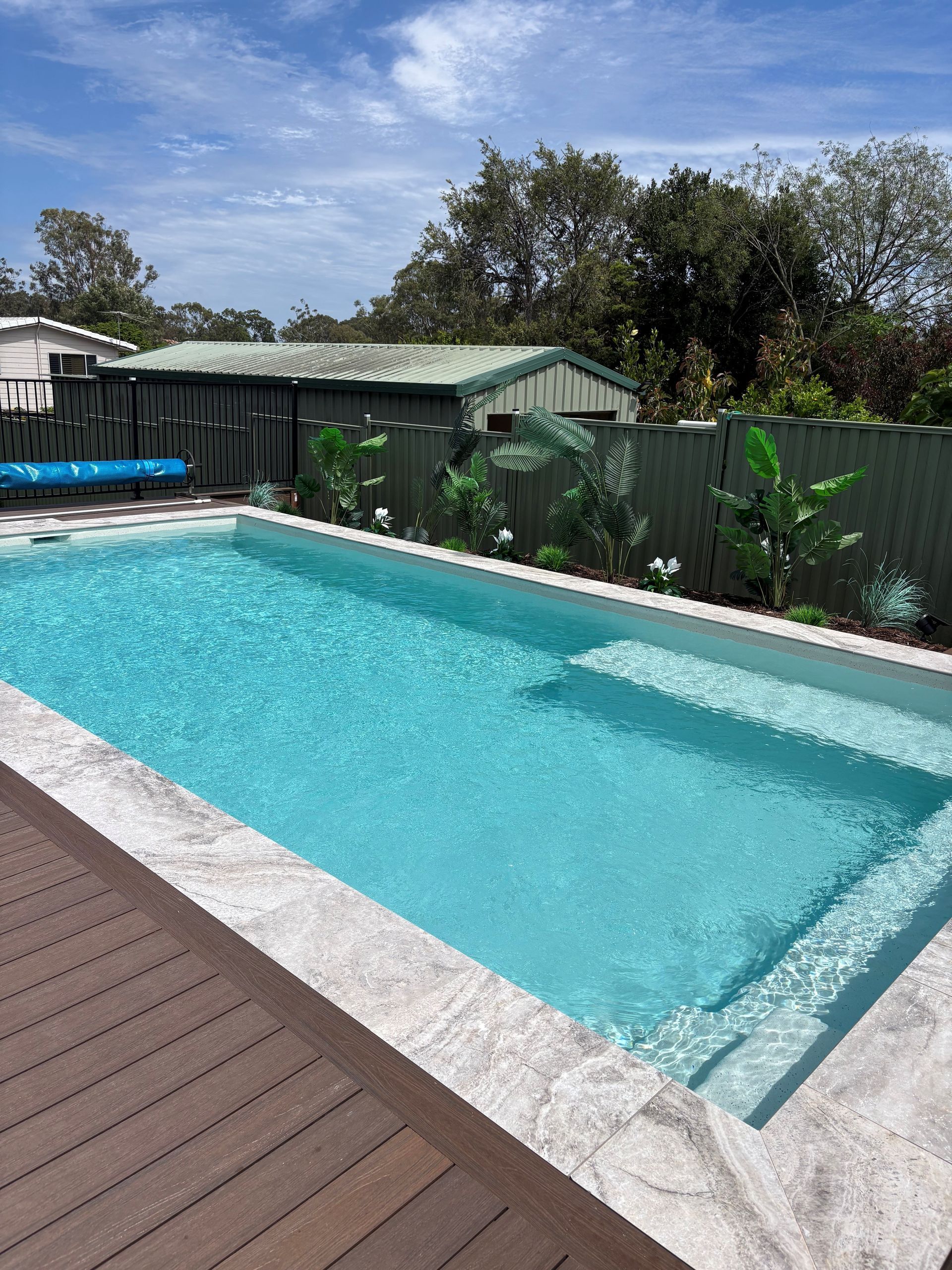 Rectangular backyard swimming pool with turquoise water, stone coping, and a wooden deck under a blue sky