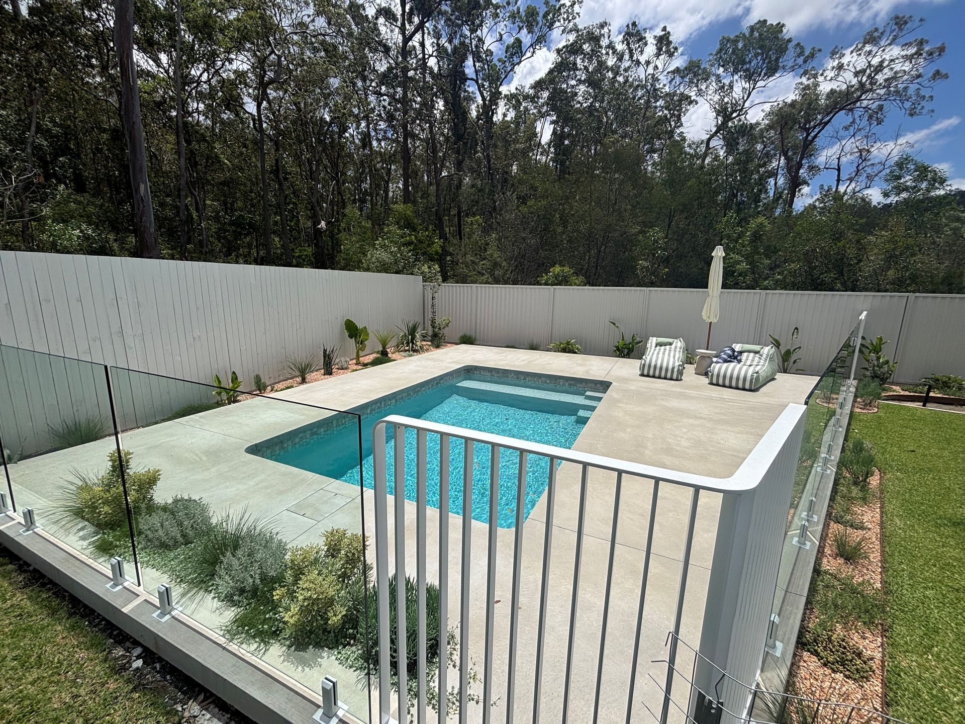 Backyard swimming pool with white fencing, patio seating, and trees beyond the wall