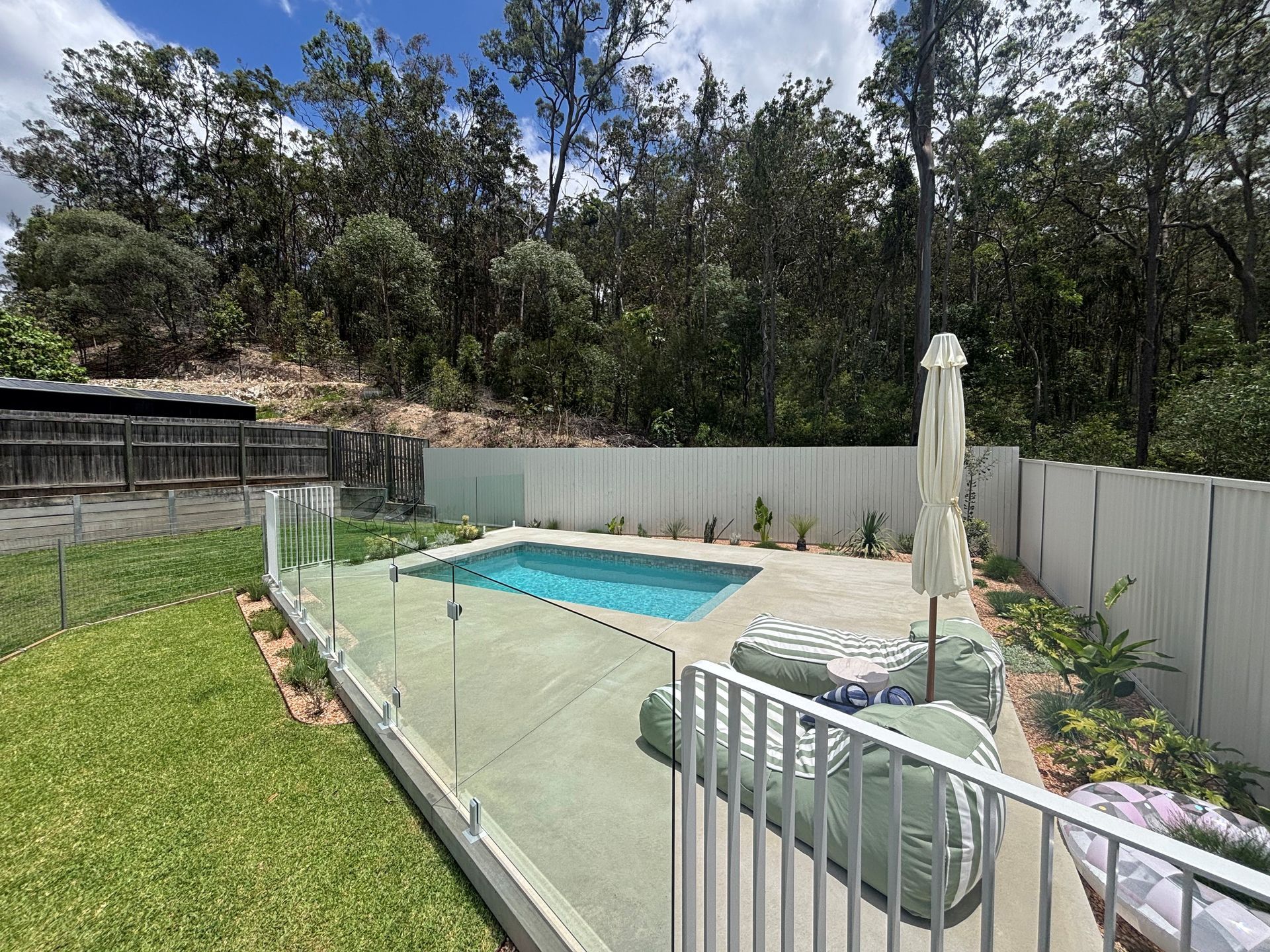 Backyard pool with glass fence, patio chairs, and white umbrella beside a grassy lawn and trees