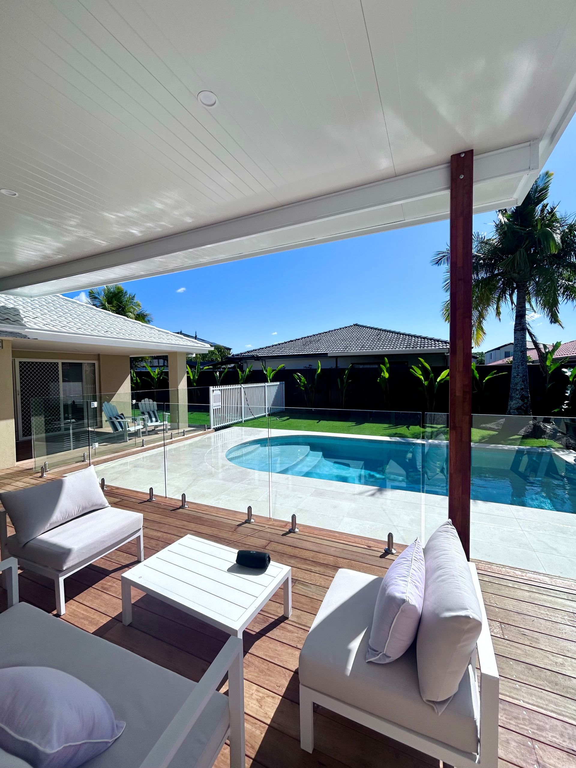 Covered patio with white seating overlooking a sunny backyard pool and umbrella-shaded lounge area