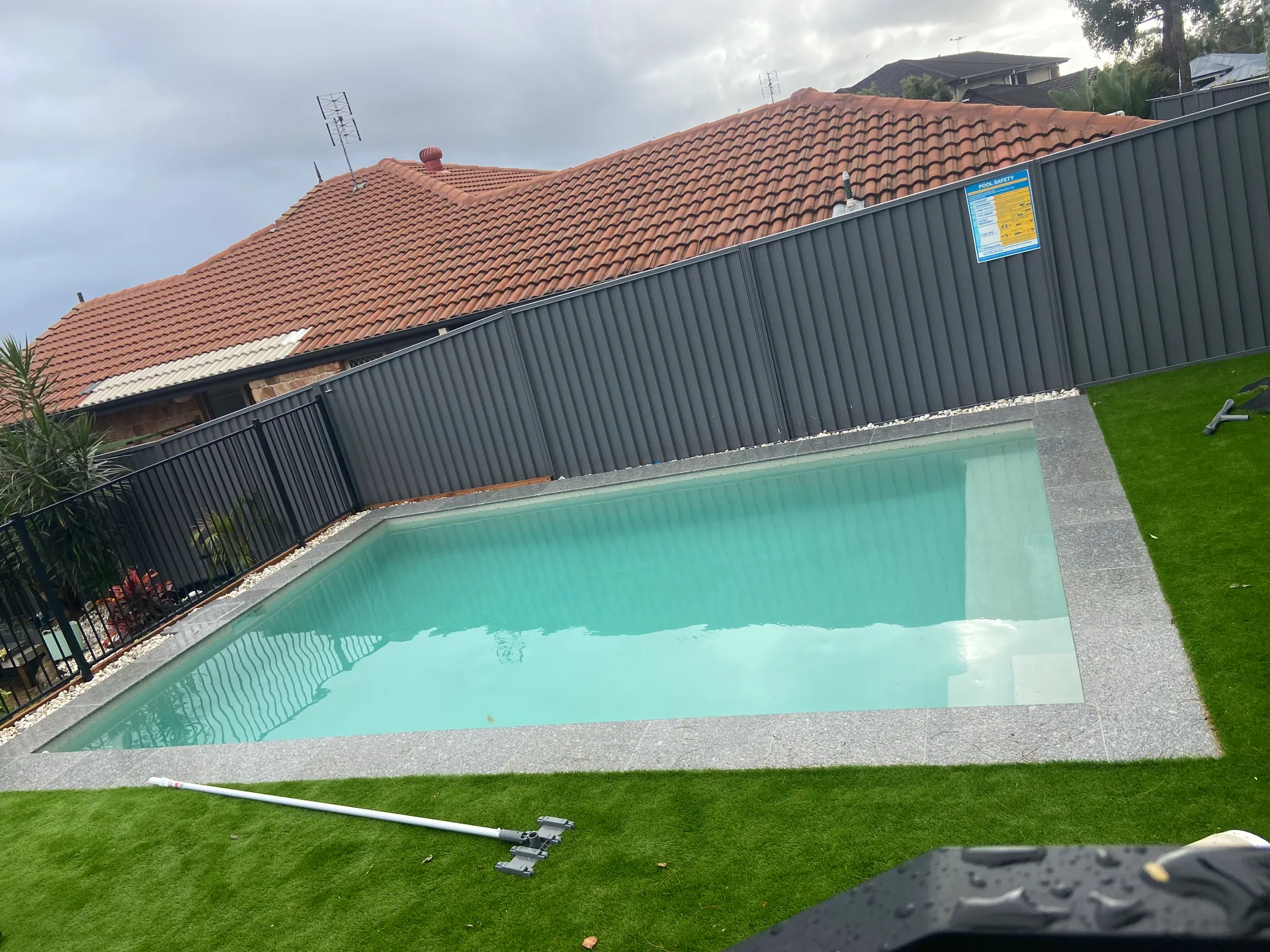 Backyard swimming pool beside a lawn and dark fence, with red-tiled roofs in the background and a skimmer pole on the grass
