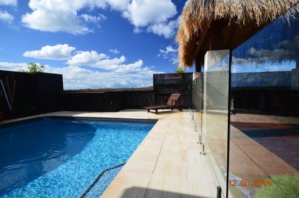 Backyard pool with blue water, white deck, glass railing, and a thatched shade against a cloudy sky