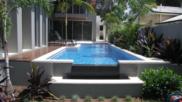 Modern backyard pool with blue water, white deck, and lush tropical plants beside a house
