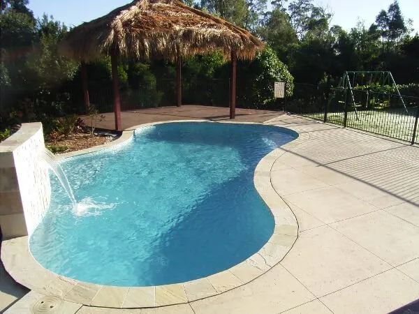Curved backyard swimming pool with blue water, beige deck, and surrounding greenery under a thatched shade structure