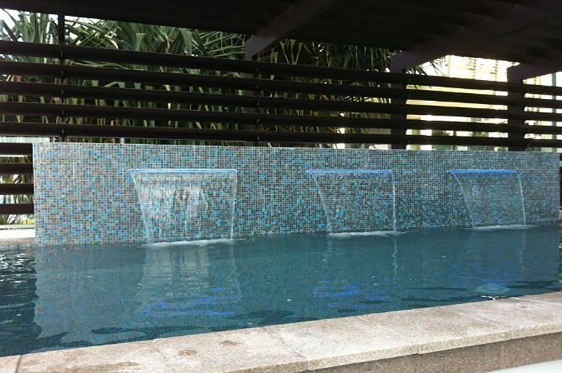 Blue-tiled backyard pool with a bubbling water wall and dark slatted pergola above