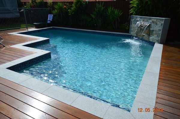 Backyard swimming pool with blue water, wooden deck, and stone water feature