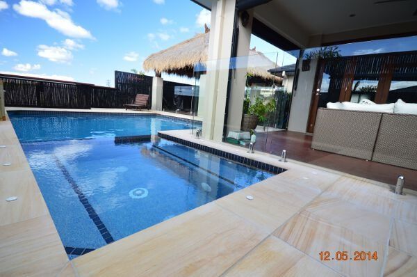 Modern backyard pool with blue water, beige deck, glass railing, and outdoor patio seating under a clear sky