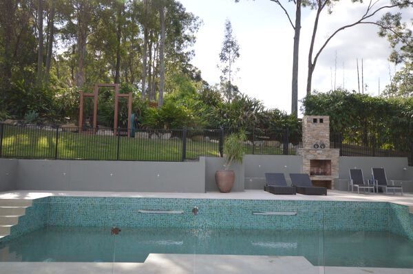 Backyard pool with tiled edge, lounge chairs, and a brick barbecue surrounded by trees and fencing