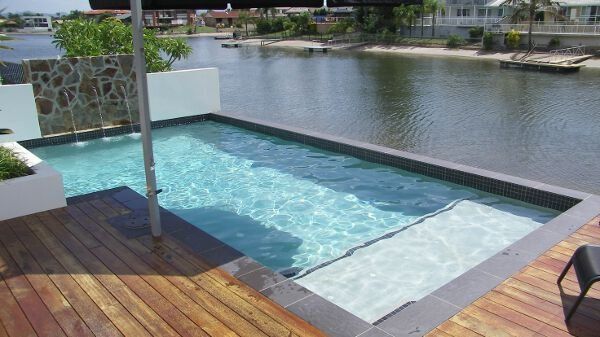 Modern backyard pool beside a canal, with wood deck and white lounge area