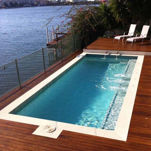 Wooden deck with a small rectangular pool beside a waterfront, with two white lounge chairs.