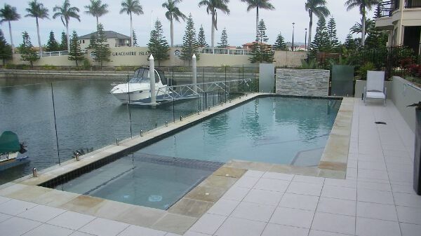 Outdoor infinity pool beside a canal, with palm trees and a sailboat in the background.