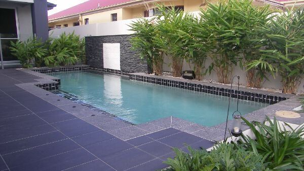 Rectangular outdoor pool with dark blue tiles, white wall fountain, and tropical plants alongside a patio