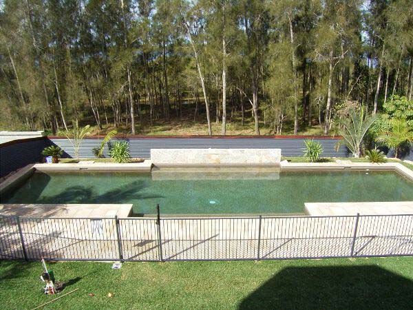 Backyard swimming pool with concrete deck, fence, and trees beyond the water.