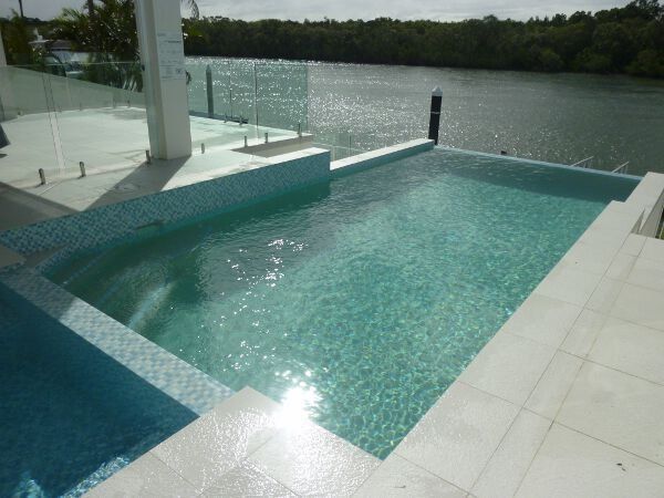 Modern pool beside a calm waterfront, with white tile decking and glass railing at sunset