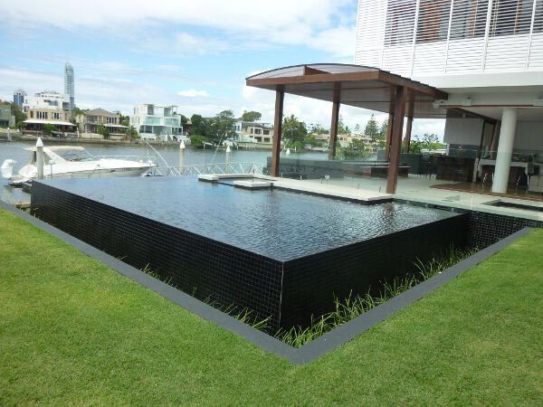 Modern waterfront patio with black reflecting pool, wooden pergola, and grassy lawn by the water.