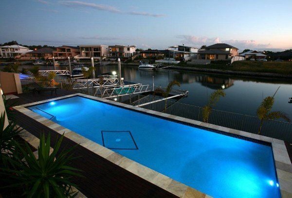 Lit blue swimming pool beside a marina and waterfront homes at dusk