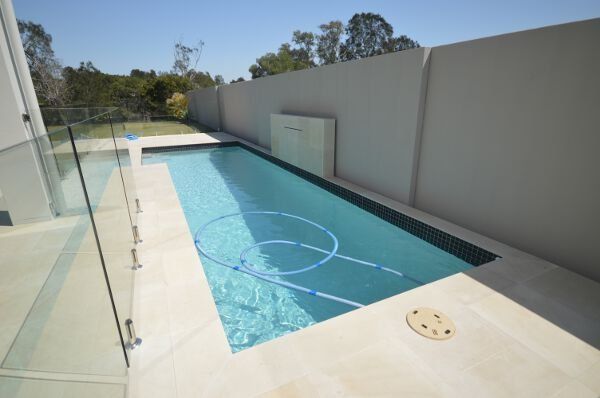 Rectangular backyard swimming pool beside a glass wall and high privacy fence under bright sunlight