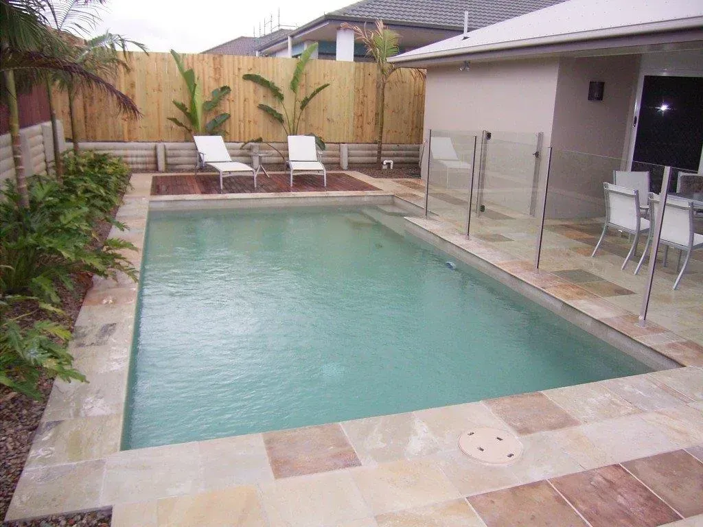 Rectangular backyard swimming pool with patio, lounge chairs, and glass fence beside a covered outdoor area