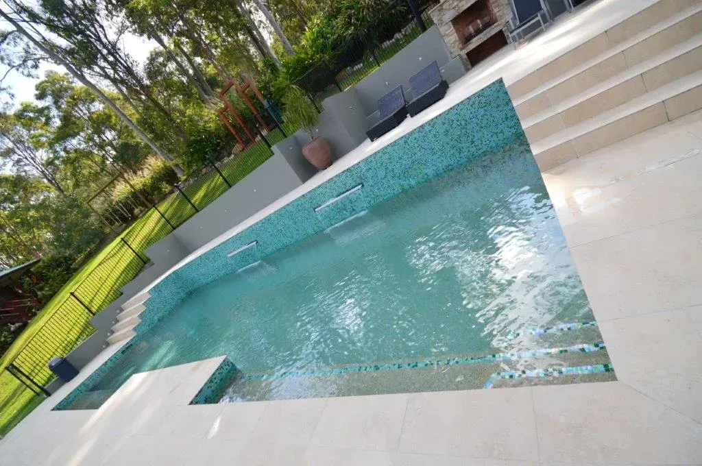Outdoor swimming pool with turquoise water, white stone deck, steps, and lounge chairs by a green lawn.