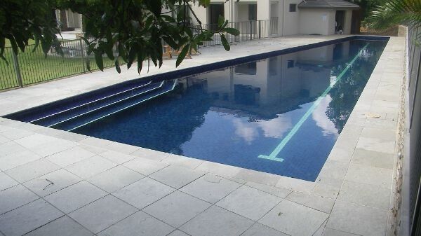 Rectangular outdoor swimming pool with dark blue water beside a light stone deck and building courtyard