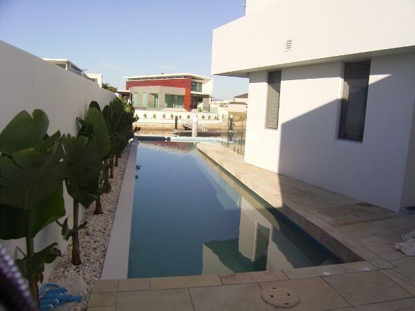 Narrow outdoor pool beside white modern buildings, with potted plants and a red building in the background