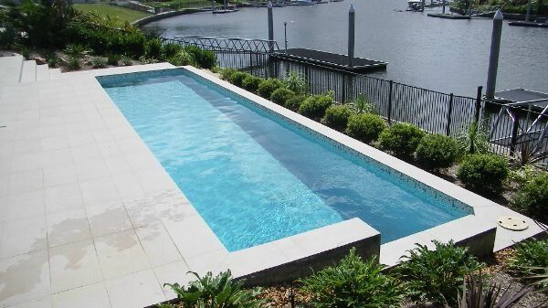 Rectangular turquoise pool beside a waterfront, bordered by white tile and greenery