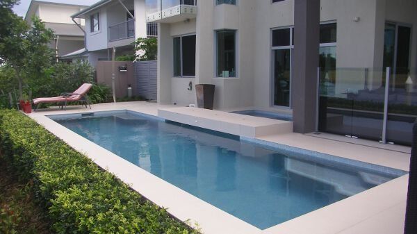 Modern house with a narrow blue swimming pool and lounge chairs beside a hedge-lined patio.