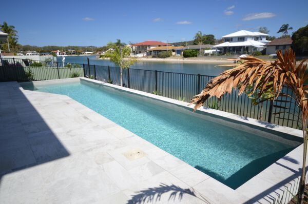 Modern backyard infinity pool with clear blue water beside a waterfront railing and houses in the distance