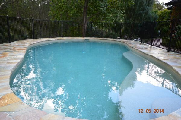 Blue swimming pool with curved edge, surrounded by trees and black fence on a stone patio.