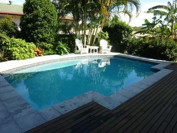 Backyard swimming pool with blue water, white patio chairs, and lush tropical plants