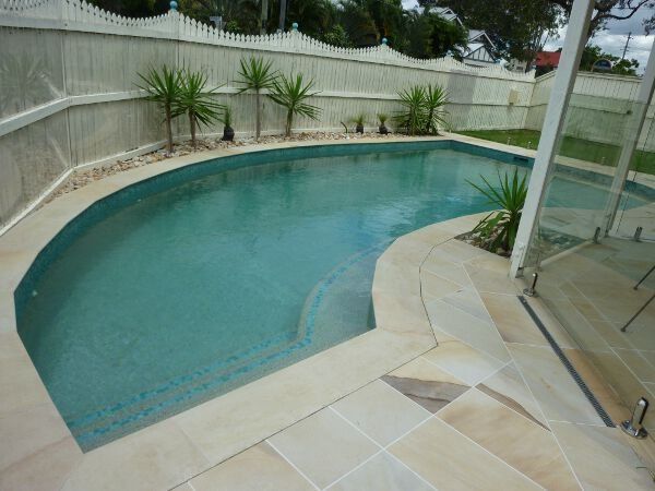 Curved backyard swimming pool with beige stone deck and potted plants beside a glass fence