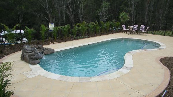 Backyard swimming pool with tan deck, rock waterfall, and lounge chairs by greenery