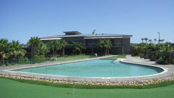 Curved outdoor pool beside a modern building, surrounded by palm trees and green lawn under a clear sky