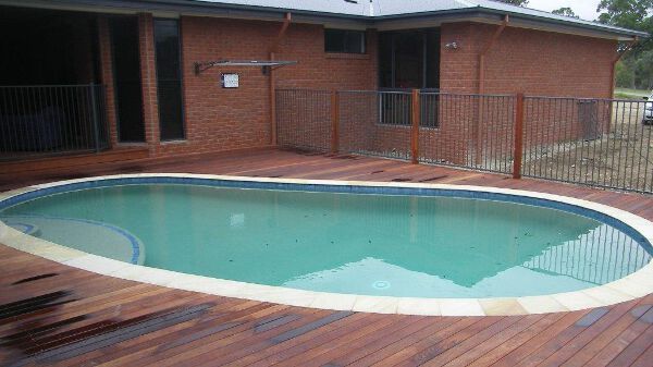 Backyard swimming pool beside a brick house, with a wooden deck and metal fence.