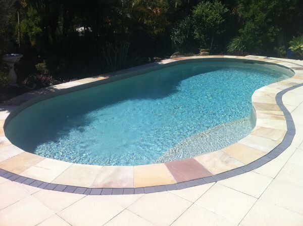 Curved outdoor swimming pool with blue water, pale stone deck, and trees in the background