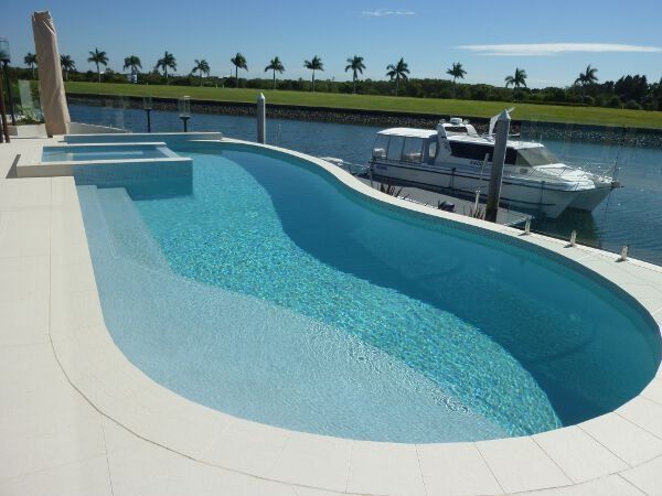 Curved blue infinity pool beside a docked white boat on a sunny waterfront patio