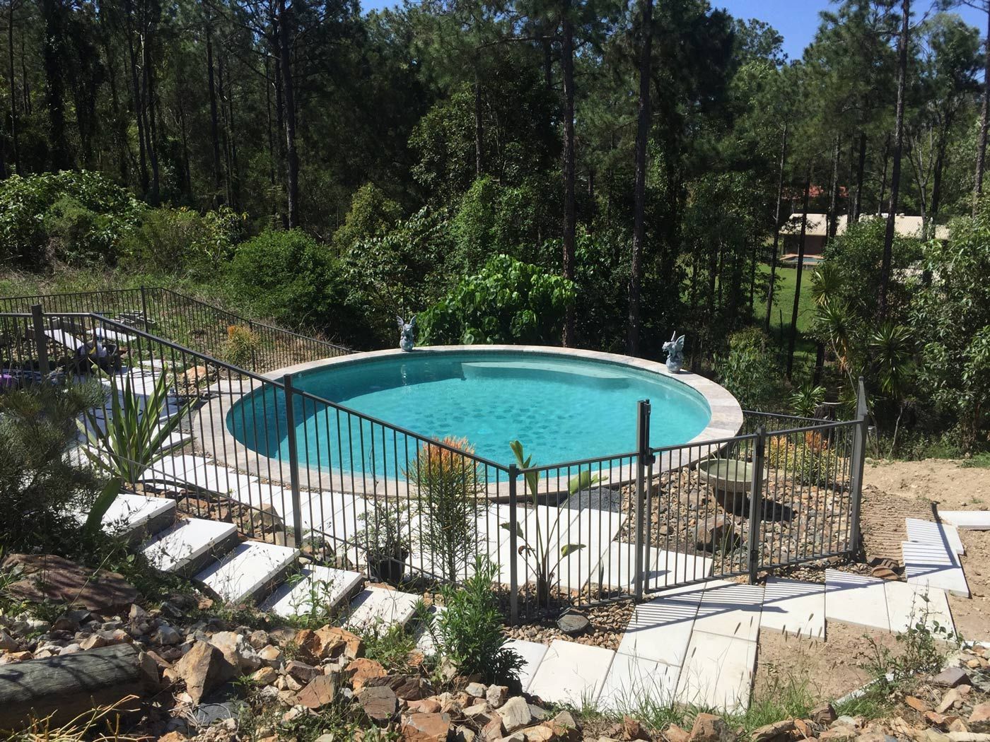 Round backyard pool surrounded by a fence and patio, with trees and rocks in the landscaped yard.