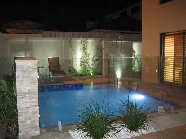 Lit backyard pool at night with lounge chair, stone pillar, and glass fence.