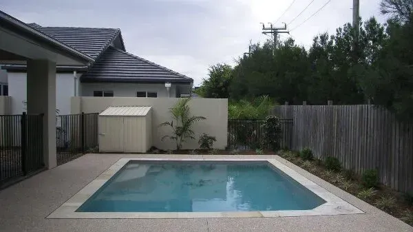 Backyard swimming pool beside a house, surrounded by a fence and trees under an overcast sky