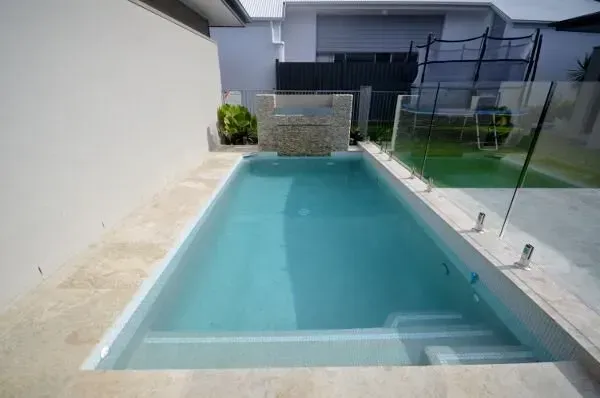 Modern backyard pool with clear blue water, glass fencing, and a small stone feature wall